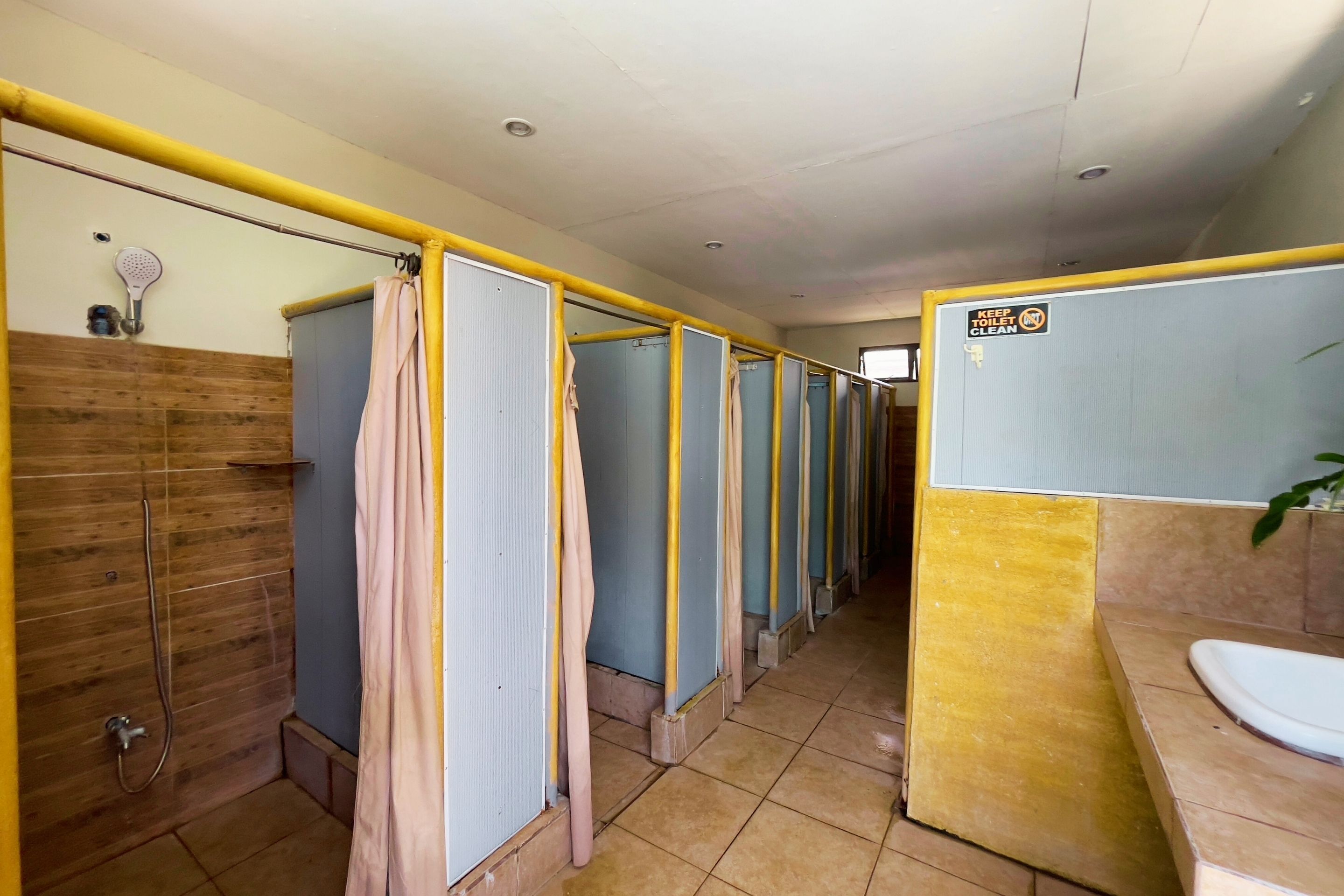 A row of individual shower cubicles with a wooden wall and a yellow partition on one side, a tiled floor, and a sink with a mirror on the opposite wall.