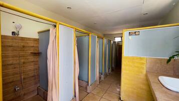 A row of individual shower cubicles with a wooden wall and a yellow partition on one side, a tiled floor, and a sink with a mirror on the opposite wall.