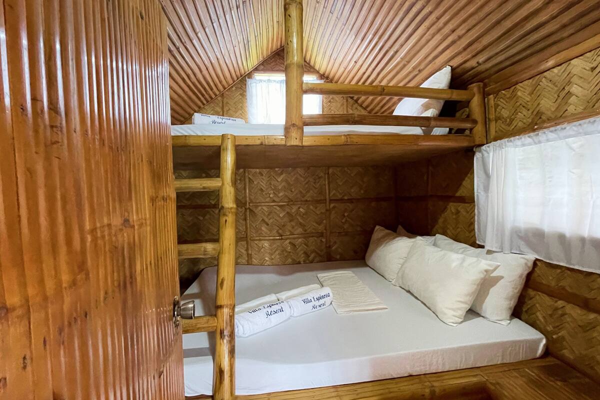 A bunk bed room with a wooden ceiling and walls, a mattress with pillows, and a window with a white curtain.