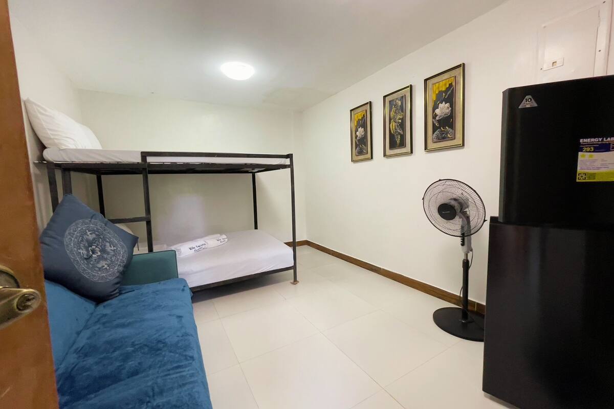 A dormitory room with bunk beds, a blue couch, a standing fan, and a refrigerator.