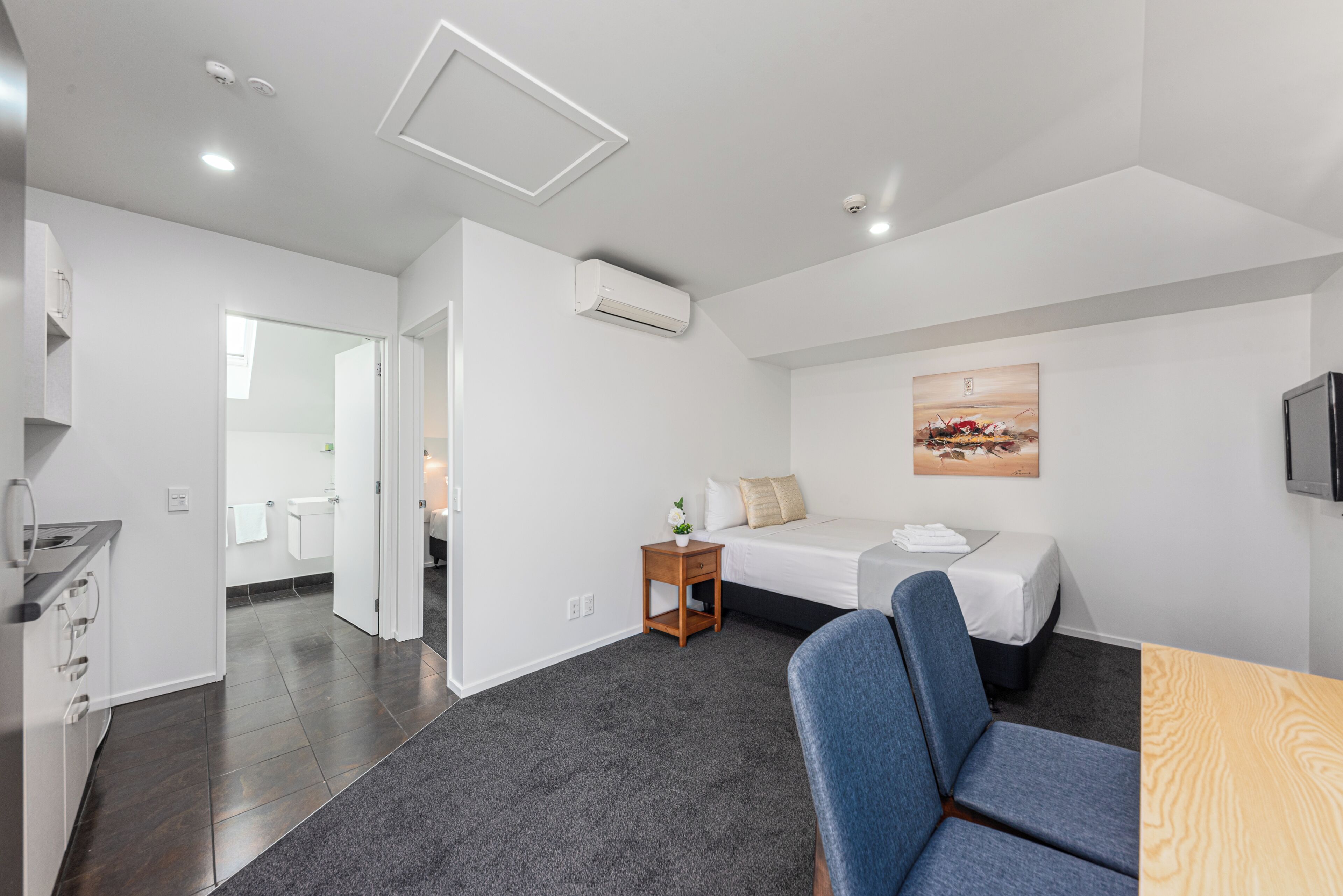 A modern hotel room with a bed, a desk, a chair, a small table with a plant, and a painting on the wall.