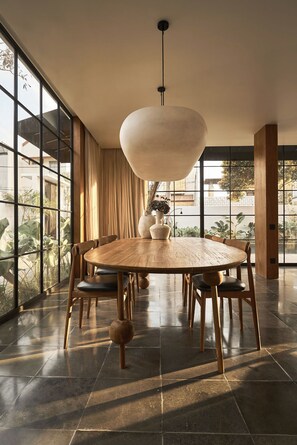 Un comedor con una mesa redonda de madera, seis sillas, una lámpara colgante y ventanas amplias con cortinas.