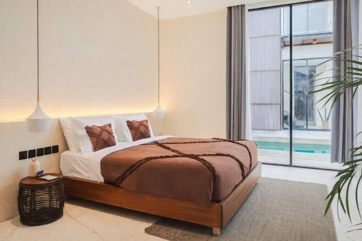 Quarto moderno com cama grande, luminárias de cabeceira, mesa de apoio e vista para uma piscina.