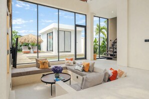 A modern living room with a large window, a sofa, a coffee table, and a view of a pool and outdoor area. - Aquamarine II By BREIG (Canggu)