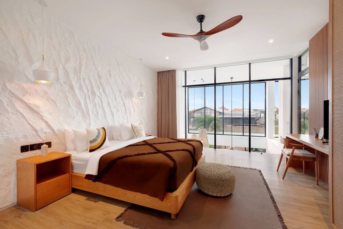 Quarto moderno com uma cama grande, uma mesinha de cabeceira de madeira, uma escrivaninha e vista para o exterior através de amplas janelas.