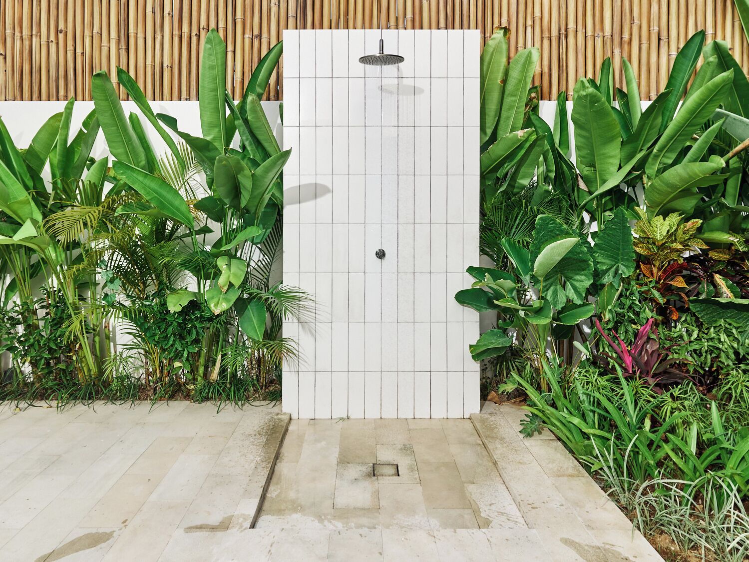 Cabina doccia in piastrelle bianche con pannello decorativo in bambù, immersa in rigogliosa vegetazione tropicale.