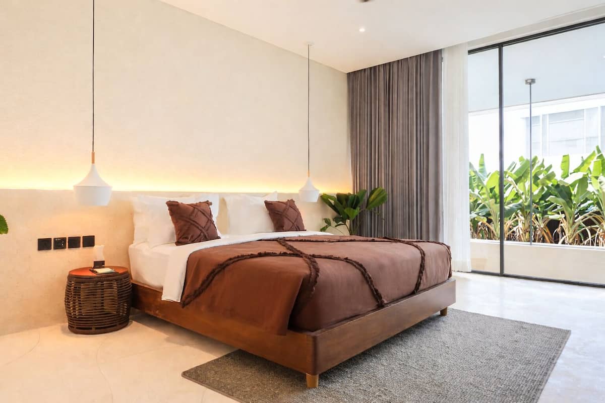 Quarto moderno com uma cama grande, luminárias de cabeceira, uma pequena mesa de apoio e vista para o verde através de uma janela.