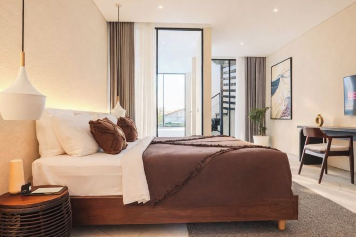 A modern hotel room with a bed, bedside table, lamp, and a view of the outside.