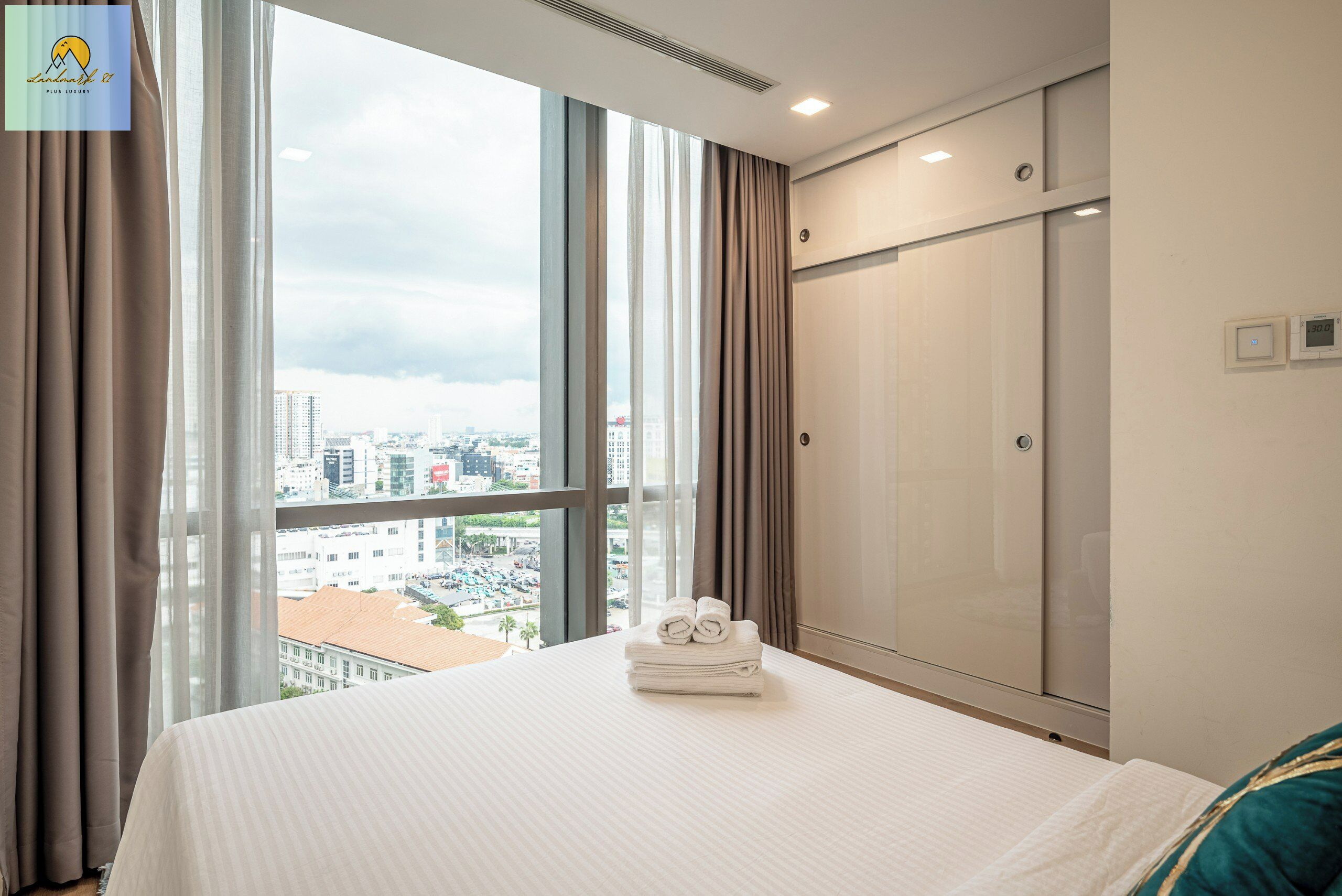 A hotel room with a large window, a bed with neatly folded towels, a wardrobe, and a view of the cityscape.