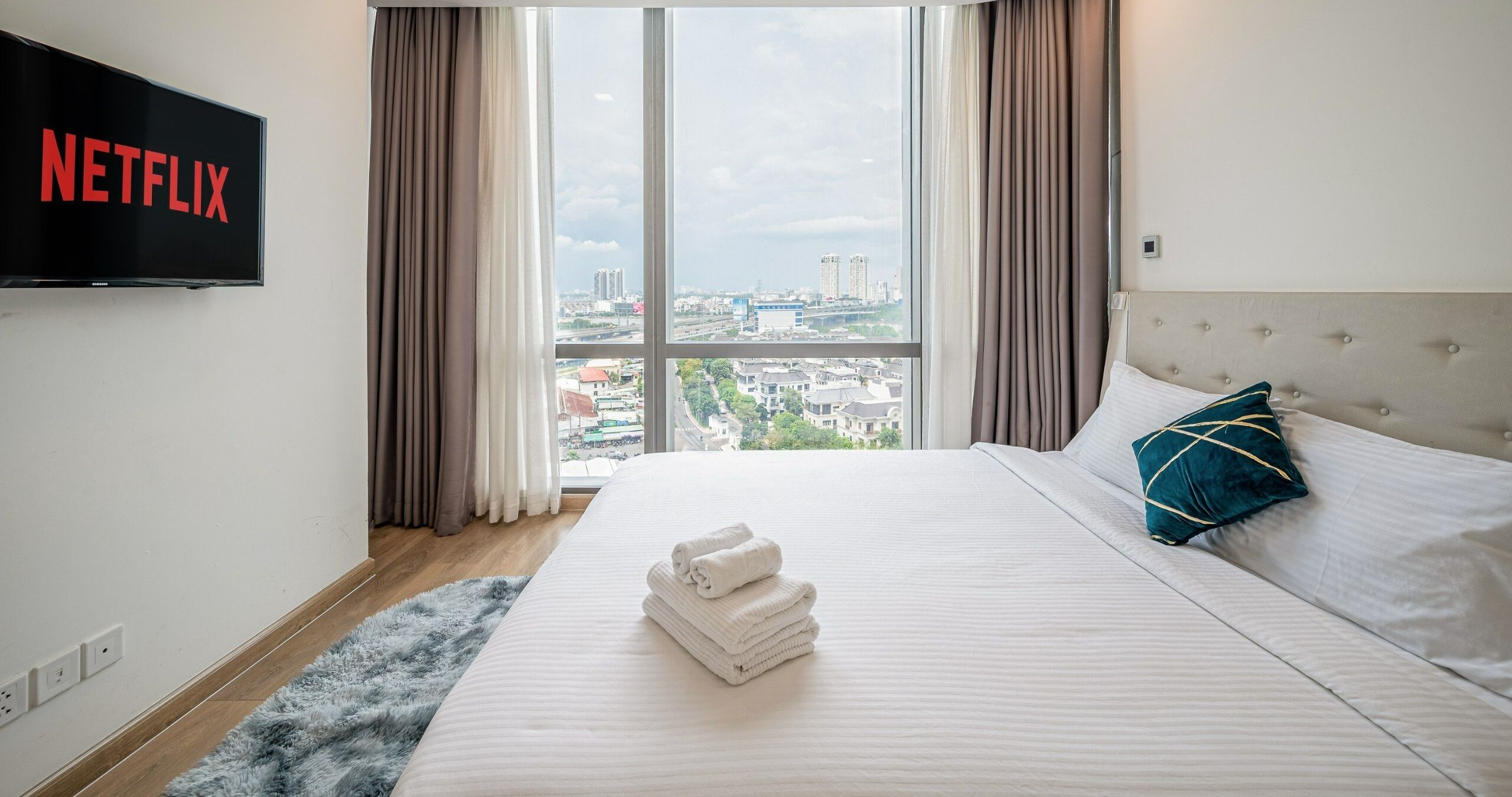 A hotel room with a large bed, a view of the city, a flat-screen TV displaying the Netflix logo, and a neatly made bed with towels.