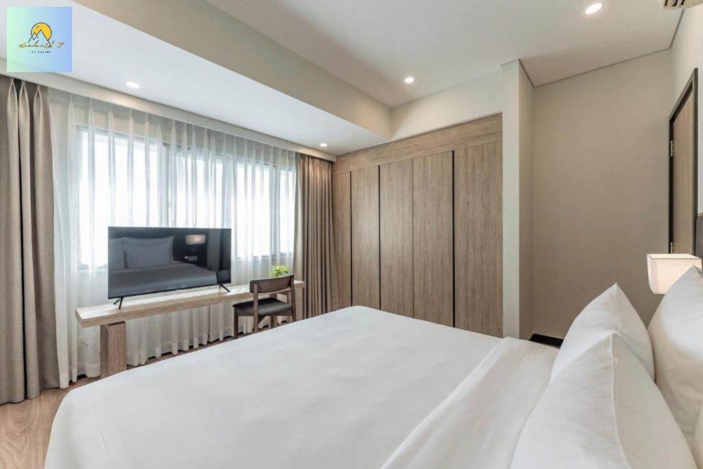 A modern hotel room with a large bed, a desk with a chair, a television, and a window with curtains.
