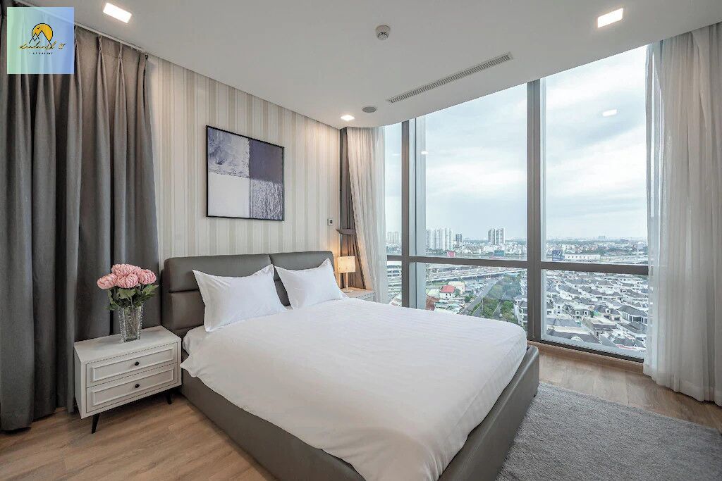 A modern hotel room with a large bed, a nightstand, a lamp, and a city view through large windows.