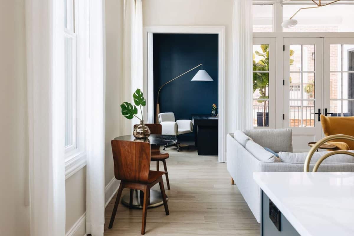 Una sala de estar moderna con un sofá, una mesa de comedor con sillas, una lámpara de pie y vistas a través de una puerta.