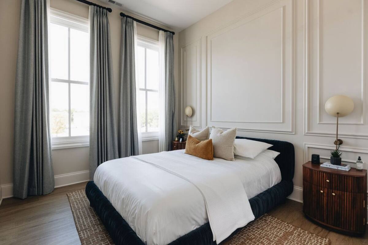 Un dormitorio con una cama grande, cortinas grises, una mesita de noche de madera y una lámpara de pared.