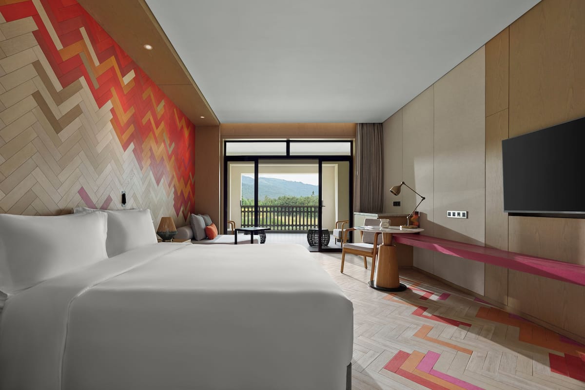 Una habitación de hotel moderna con una cama grande, un escritorio con una silla, un televisor montado en la pared y un balcón con vistas a las montañas.
