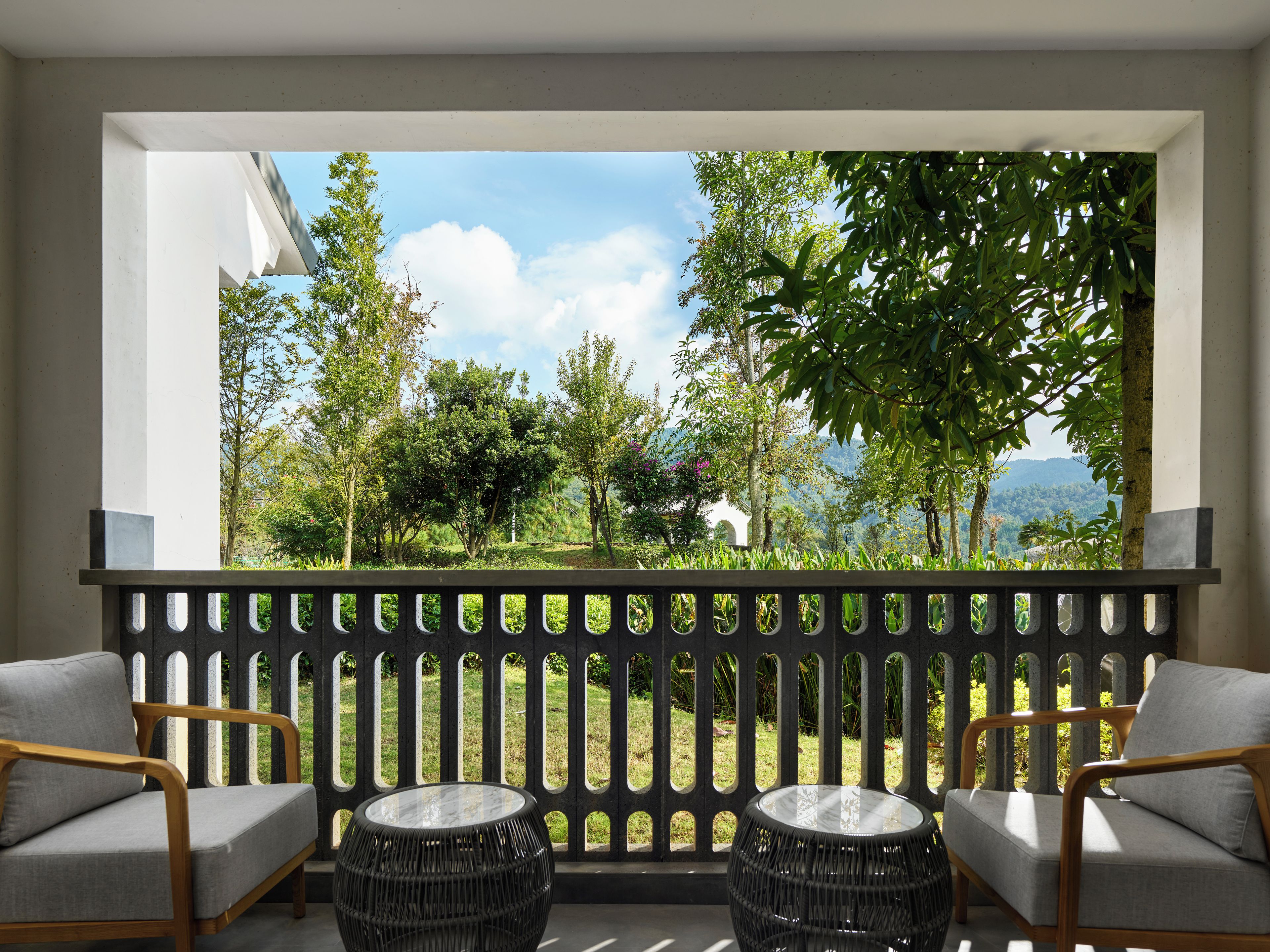 A balcony with two chairs and two round tables, overlooking a garden with trees and a mountain view.
