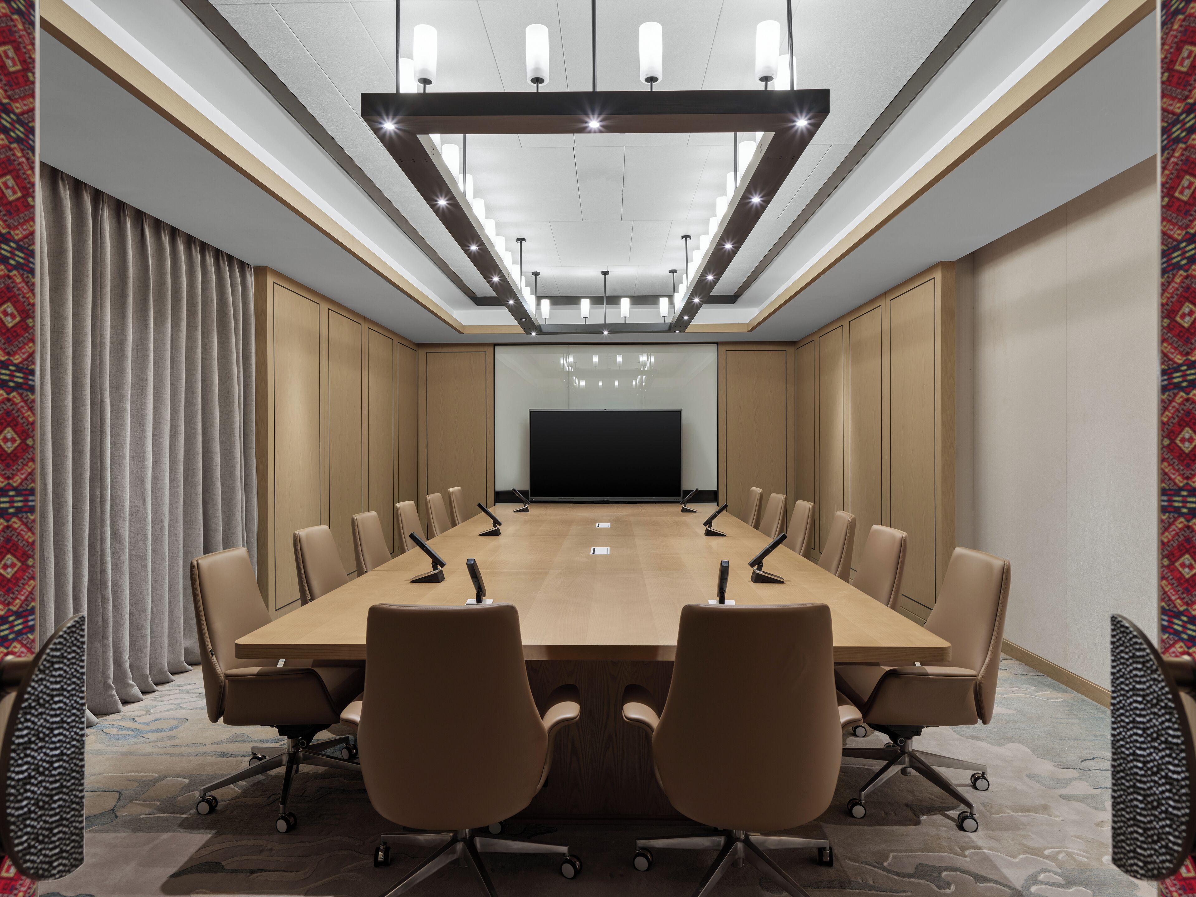A conference room with a long table, multiple chairs, a projector screen, and a modern ceiling light fixture.