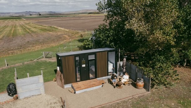 Un piccolo e moderno chalet con facciata in legno e ampie finestre, immerso nella campagna con vista su campi aperti e un albero nelle vicinanze.