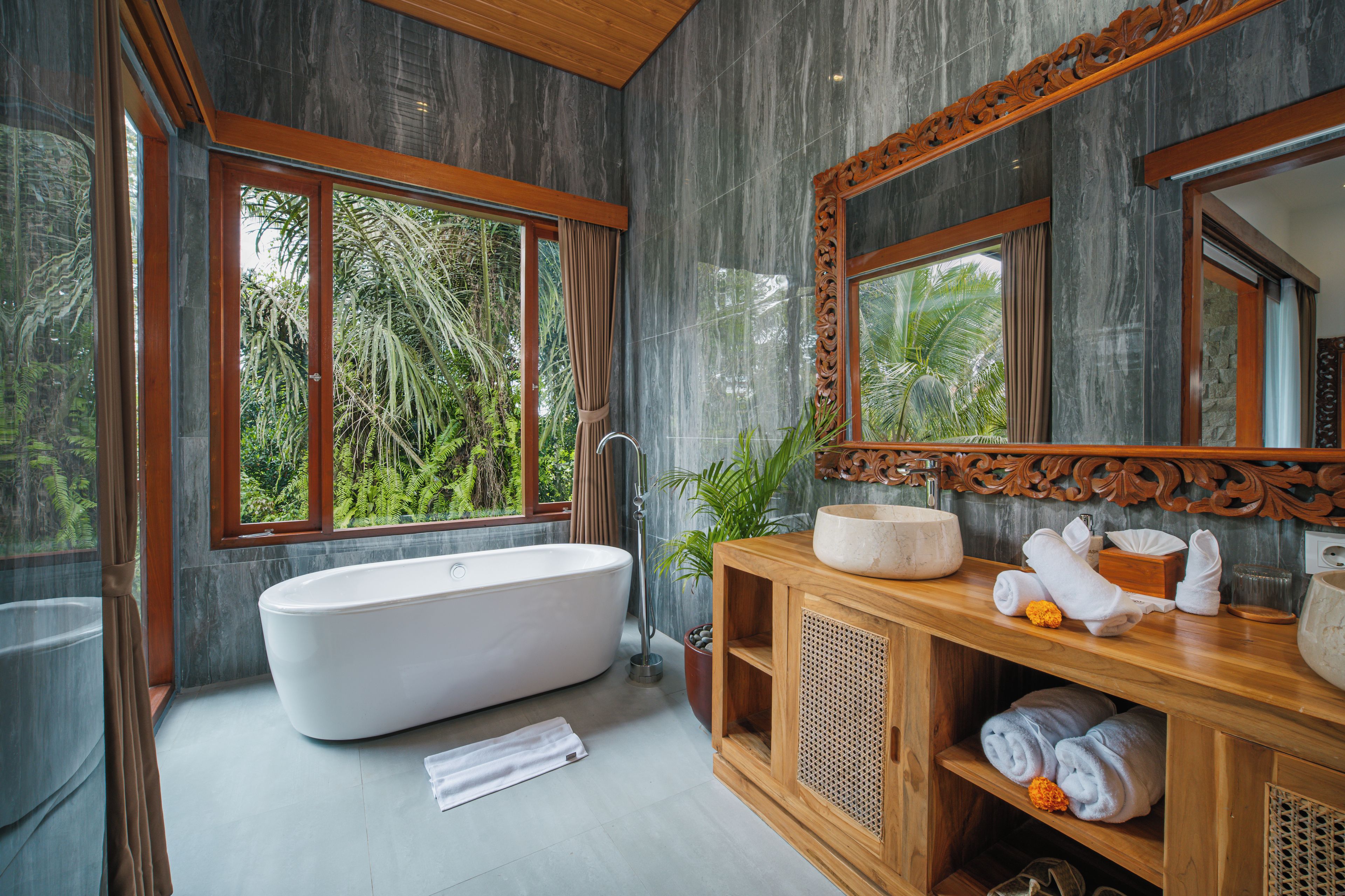A modern bathroom with a freestanding bathtub, wooden vanity, and large windows offering a view of lush greenery.