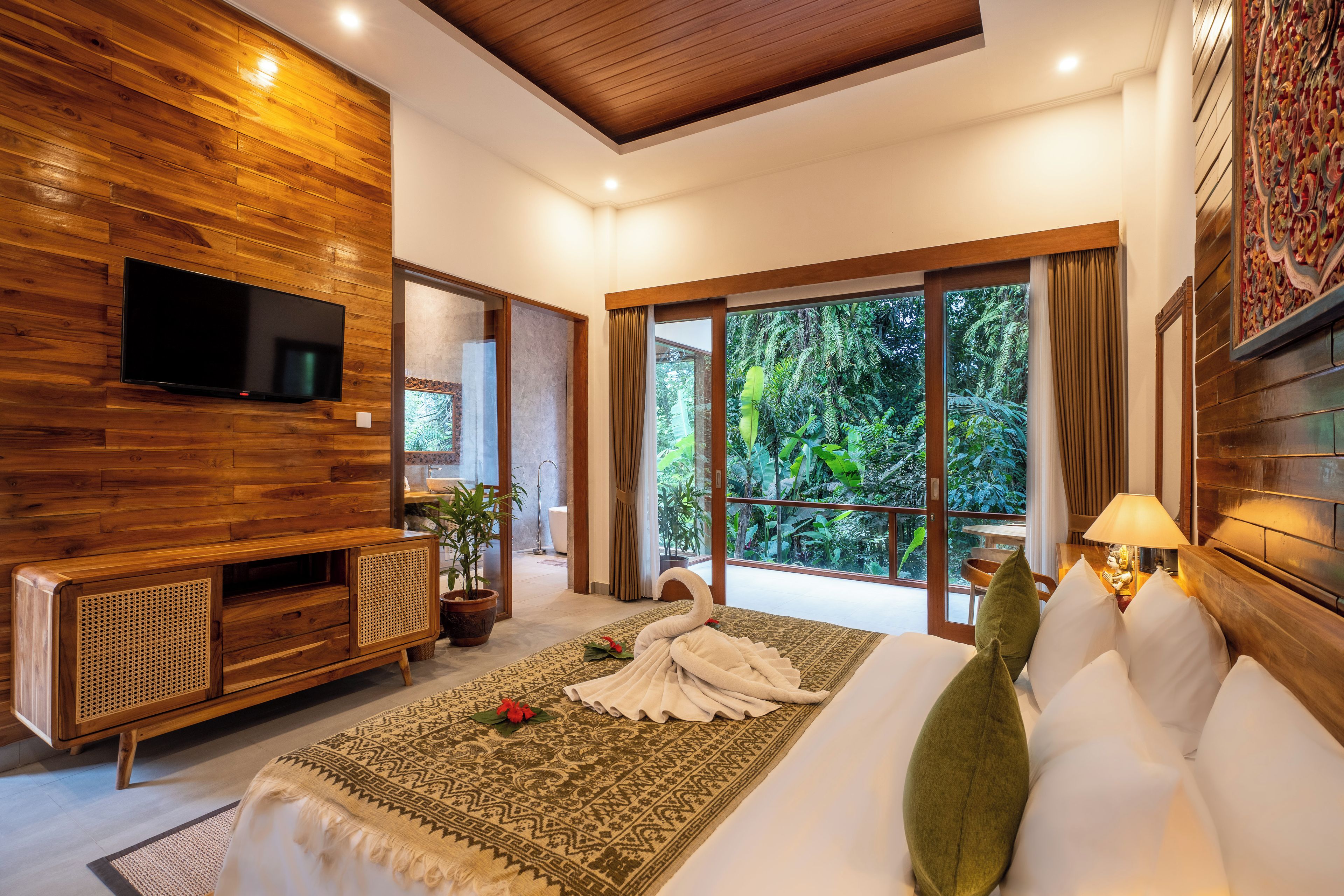 Un dormitorio moderno con pared de madera, televisor de pantalla plana, sofá, cama con cabecera decorativa y vista a follaje exuberante a través de grandes ventanales.
