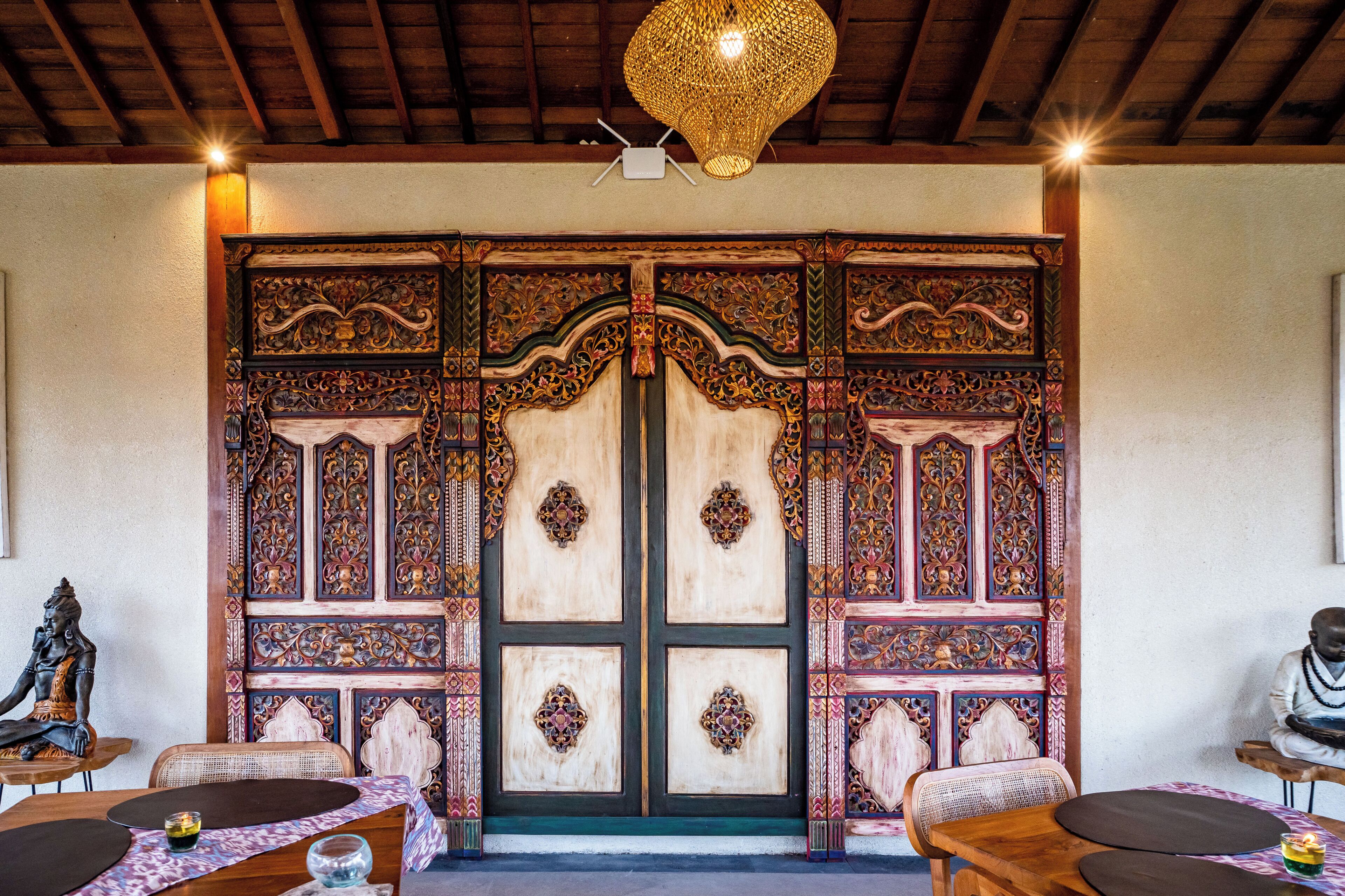 A traditional wooden door with intricate carvings, a woven ceiling light, and a statue of a deity on the left.