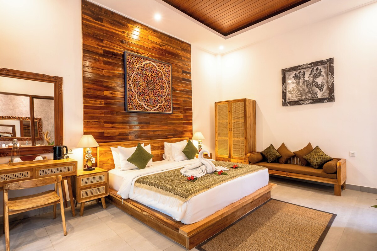 Quarto de hotel com cabeceira de madeira, uma cama grande com roupa de cama branca, mesa de cabeceira de madeira, um banco de madeira e um armário fixado na parede.