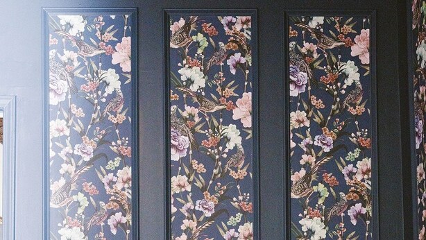 Una habitación con una pared azul oscuro, tres obras de arte florales enmarcadas verticalmente, un sillón de cuero, una silla de madera con un cojín rojo y una pequeña mesa redonda con una tetera y una figurita.