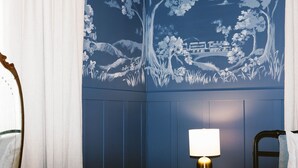 Un dormitorio con una pared azul, una mesita de noche de madera con una lámpara, y una cama con una colcha blanca.