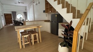 Un vaste espace de vie doté d’un plancher en bois, une table à manger centrale entourée de tabourets, une cuisine avec un réfrigérateur noir et un escalier en bois.