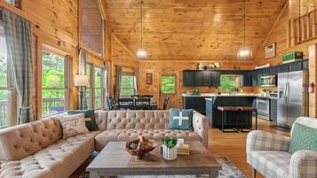 A spacious living room with a large beige sofa, a coffee table, and a view of the kitchen and dining area through large windows.