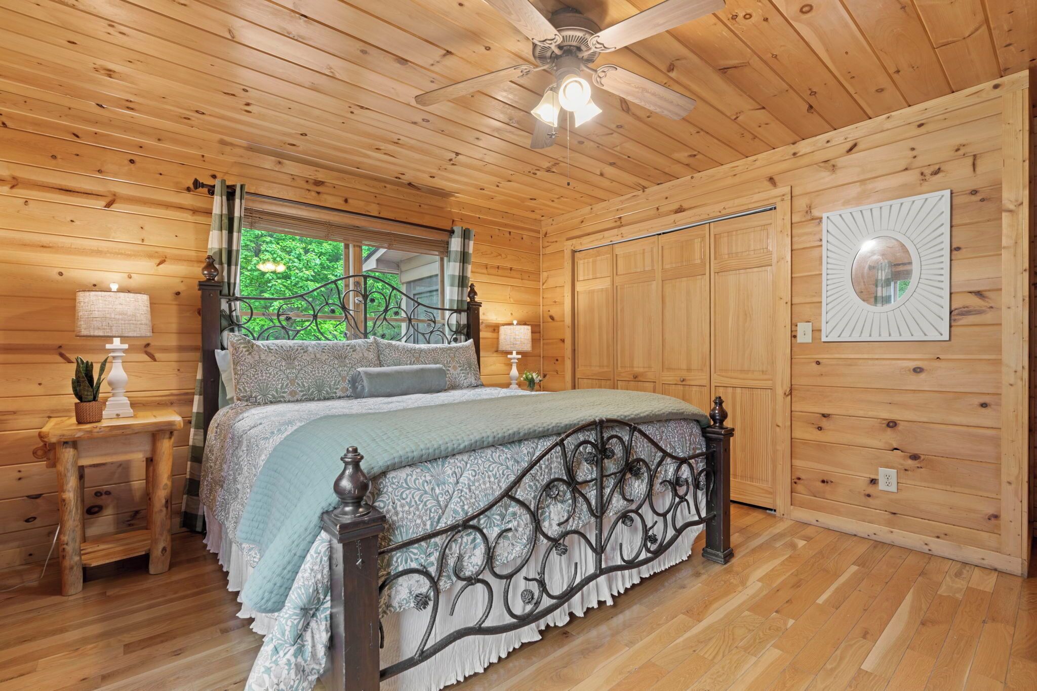 A wooden cabin bedroom with a bed, two bedside tables, a ceiling fan, and a window with a view of greenery.