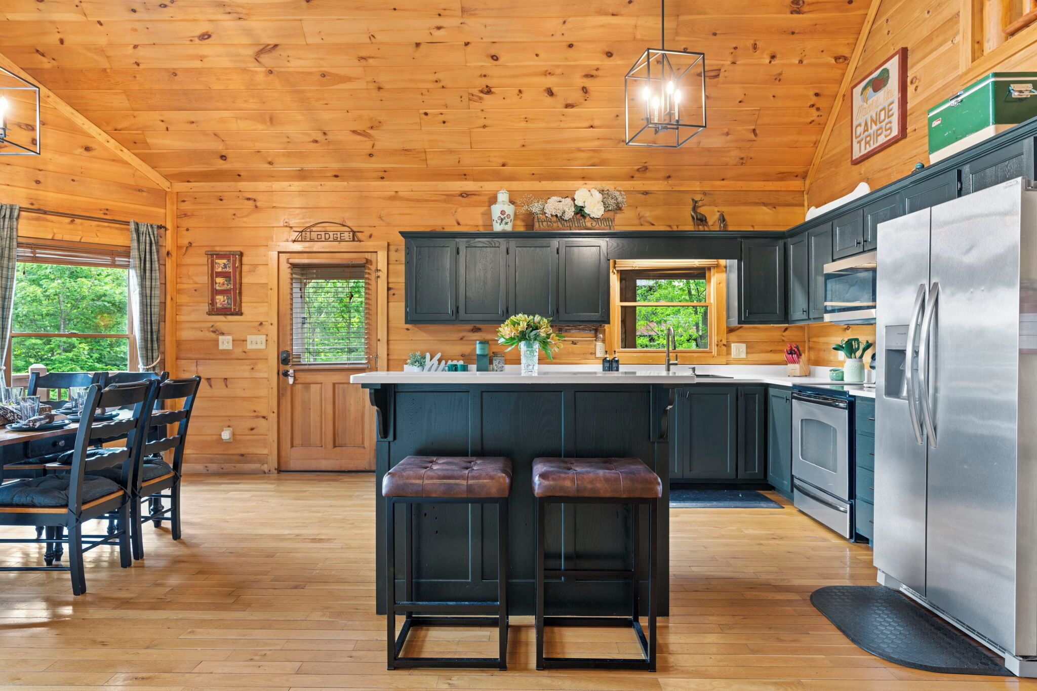 A wooden cabin with a kitchen featuring dark cabinets, a stainless steel refrigerator, and a dining area with a table and chairs.
