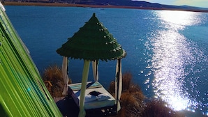 A gazebo with a thatched roof overlooking a body of water, with a clear sky and distant mountains. - Titicaca Glampina (Puno)