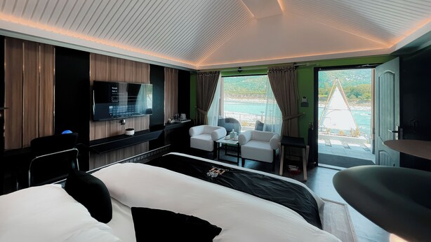 Una habitación de hotel moderna con cama, escritorio con televisor de pantalla plana, zona de estar con sillas y vista al mar a través de amplias ventanas.