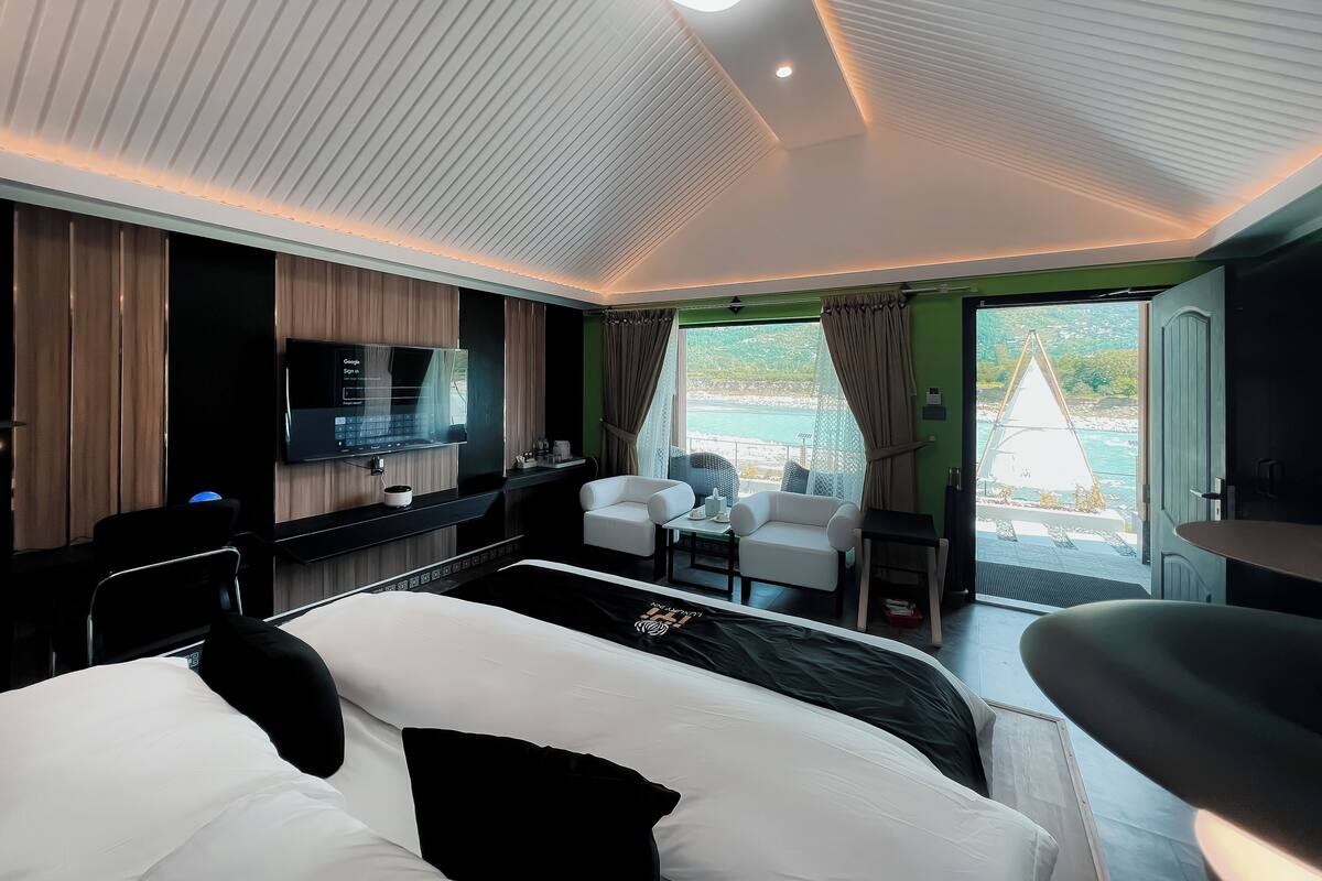 Une chambre d’hôtel moderne équipée d’un lit, d’un bureau avec un téléviseur à écran plat, d’un coin salon avec des fauteuils et offrant une vue sur la mer grâce à de grandes fenêtres.