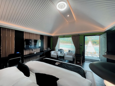 A modern hotel room with a bed, a desk with a flat-screen TV, a seating area with chairs, and a view of the sea through large windows.