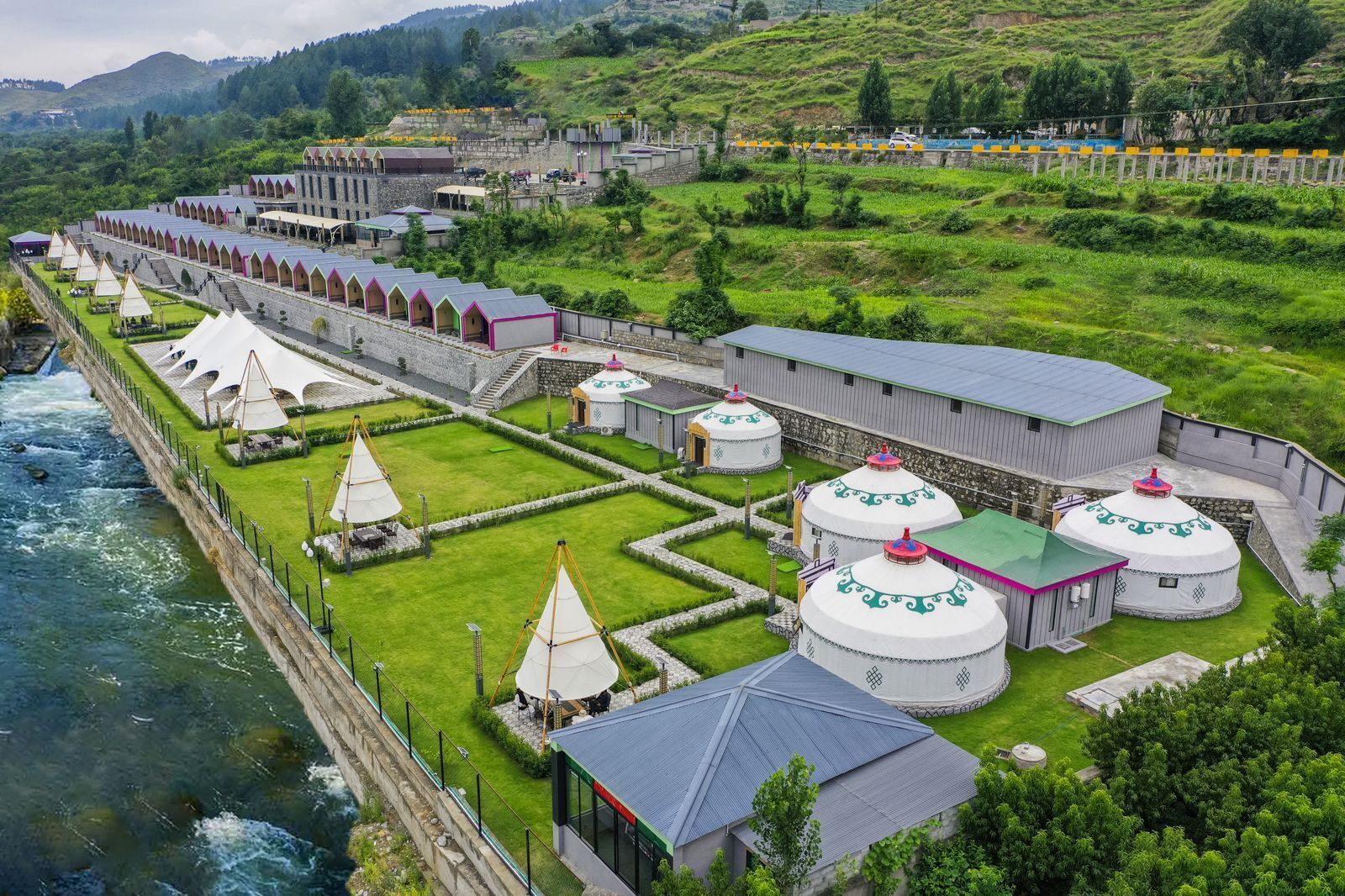 A traditional yurt-style building complex with yurts and a central structure, surrounded by a river and lush greenery.