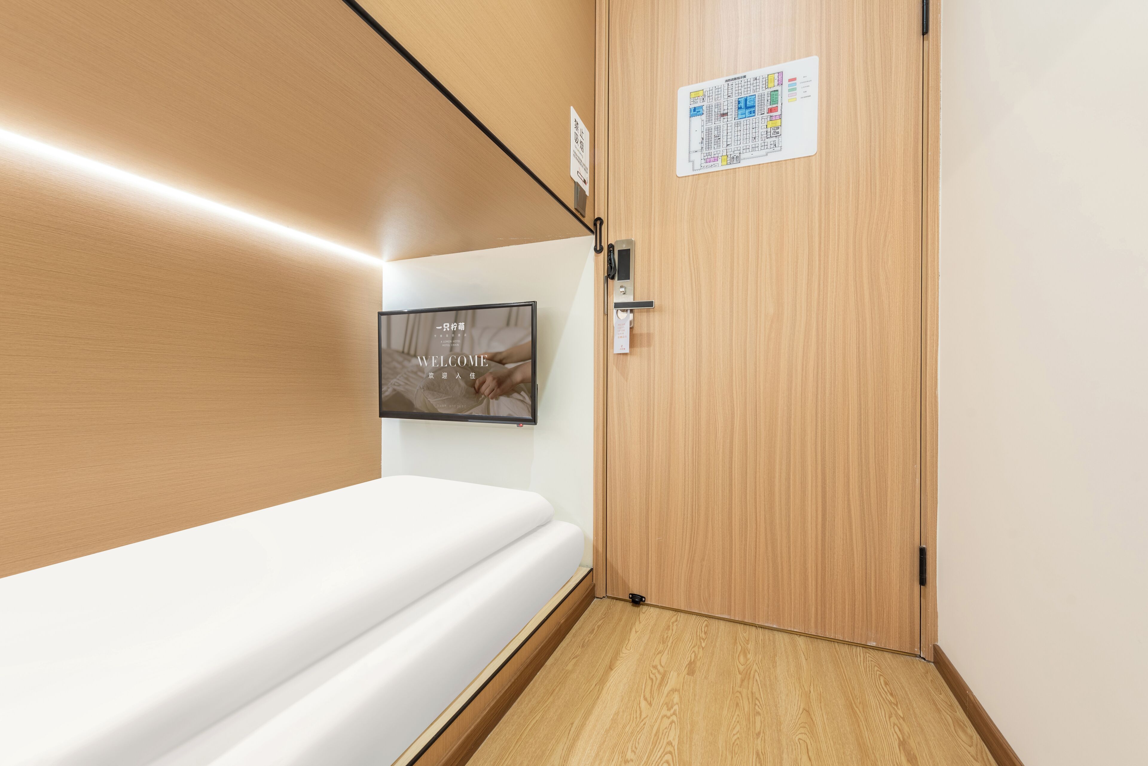 Een hotelkamer met een houten vloer, een wit bed, een aan de muur bevestigde televisie en een deur met een kaart en een bordje.