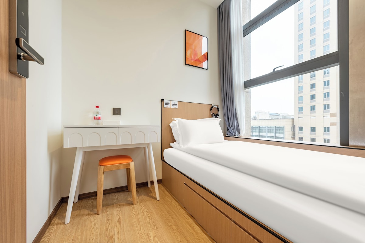 A hotel room with a bed, a desk with a chair, a window with a view of buildings, and a picture on the wall.