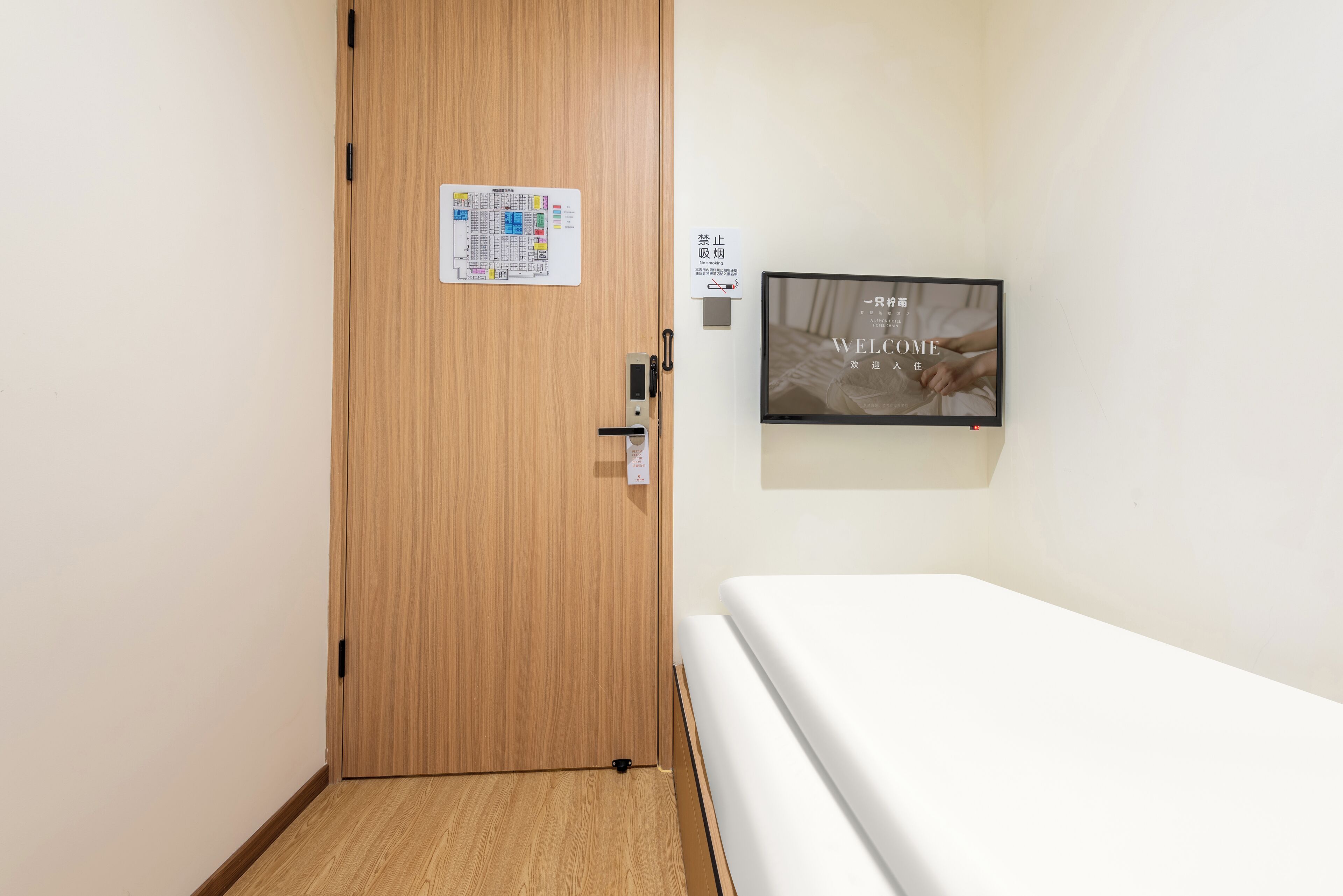 Habitación de hotel con puerta de madera, televisor empotrado mostrando 'WELCOME HOME' y cama con ropa blanca.