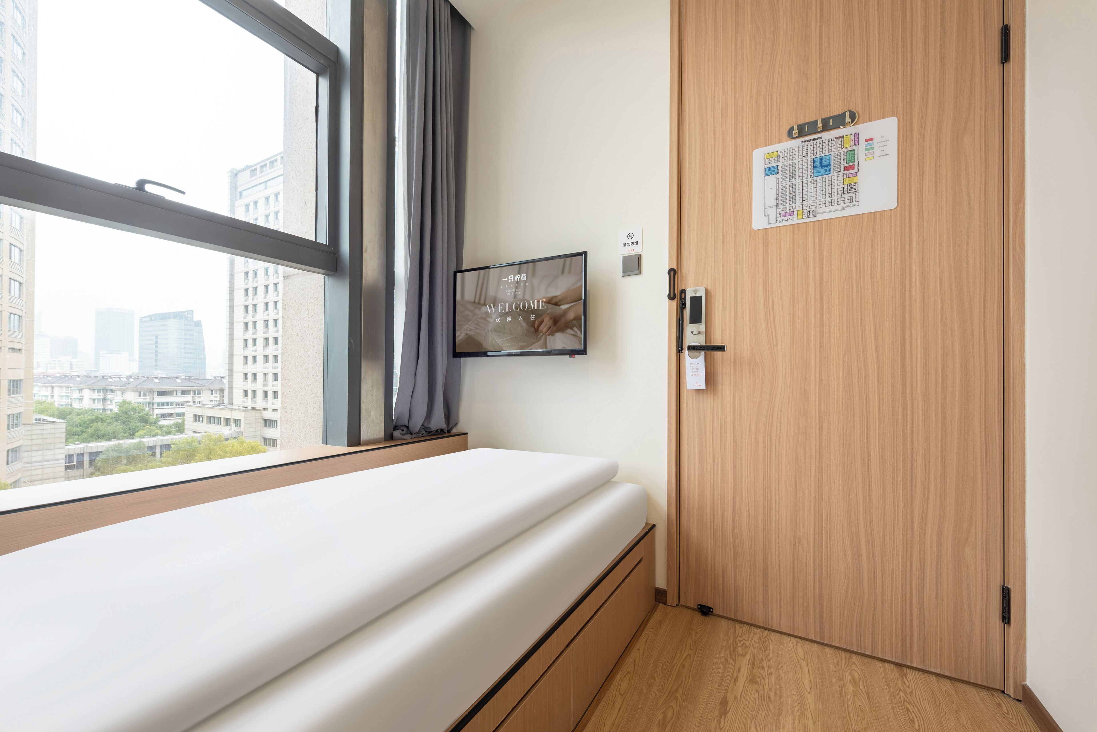 A hotel room with a bed, a window with curtains, a wall-mounted TV, a door with a lock, and a bulletin board with a welcome sign.