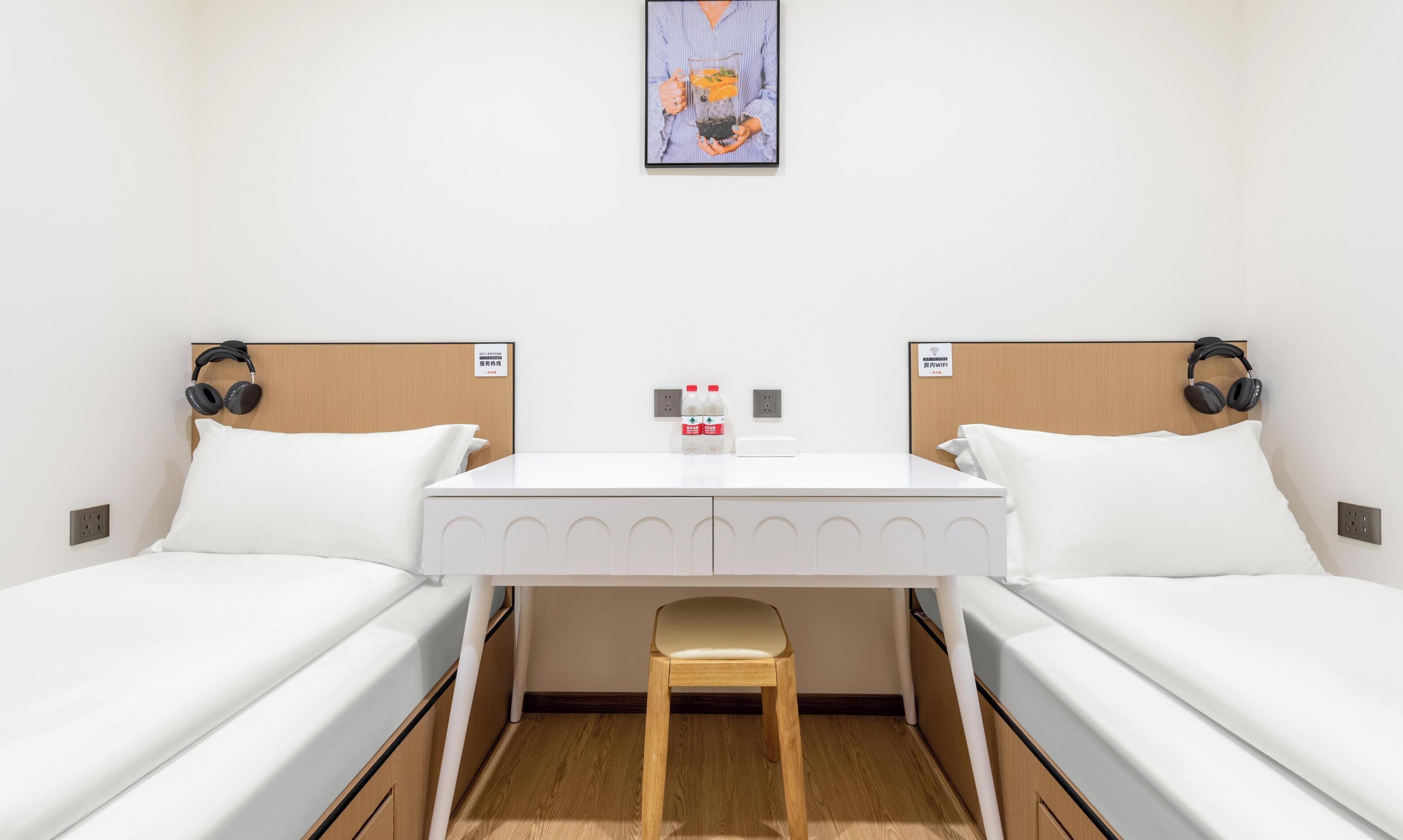 A hotel room with two single beds, a small table, a chair, and a painting on the wall.