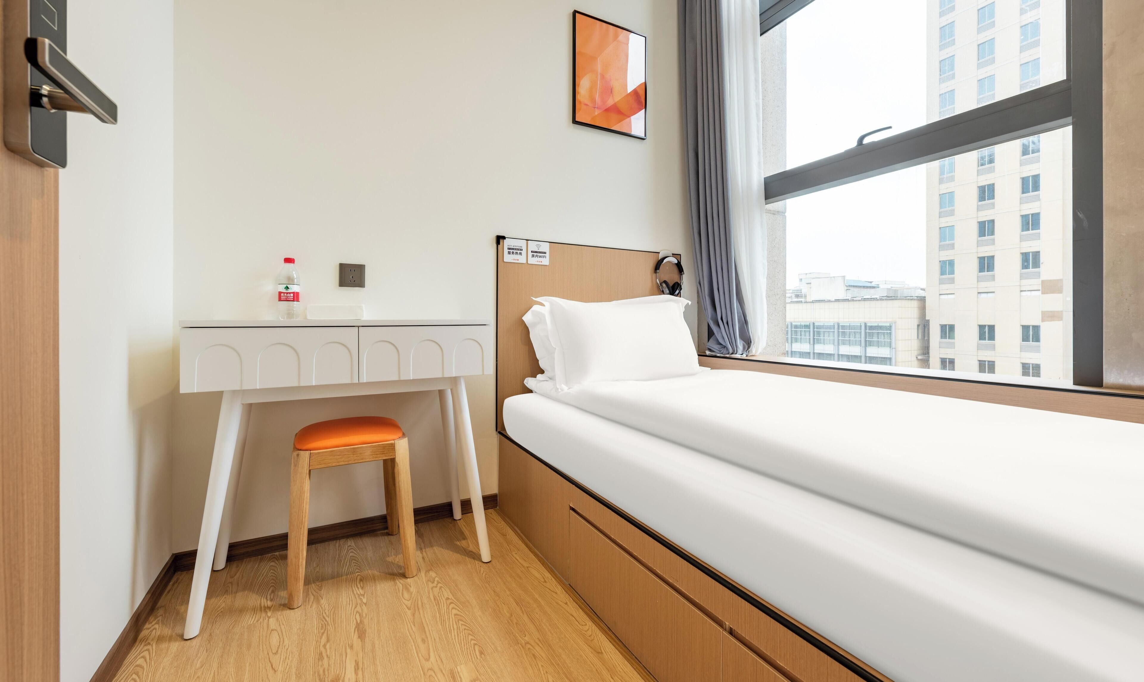 A hotel room with a bed, a desk, a chair, a window with a view of buildings, and a wall-mounted artwork.