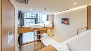 Ein kleines Hotelzimmer mit Etagenbett, einem kleinen Schreibtisch, einem Fenster mit Vorhängen und einem Bild an der Wand.