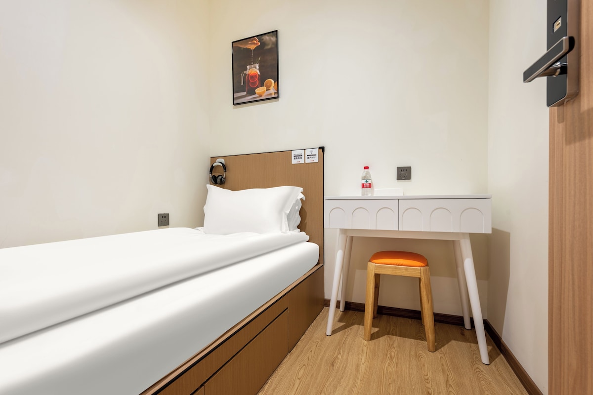 A hotel room with a single bed, a desk with a chair, and a wall-mounted shelf.