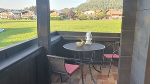 A balcony with a round table and two wicker chairs, overlooking a green field and houses. - Ferienwohnung Nr. 23, 55 qm, 2 Personen, 1 Schlafzimmer, Balkon (Unterwössen)