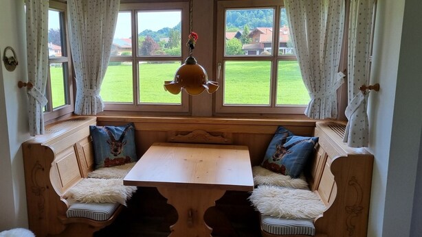 A cozy room with a wooden bench and a table, large windows with curtains, and a view of a green landscape. - Ferienwohnung Nr. 23, 55 qm, 2 Personen, 1 Schlafzimmer, Balkon (Unterwössen)