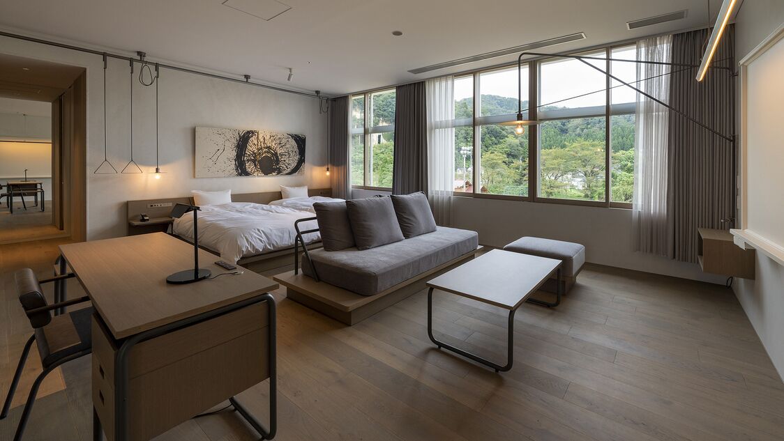 Een moderne hotelkamer met een bed, een bank, een bureau en grote ramen met uitzicht op groen.