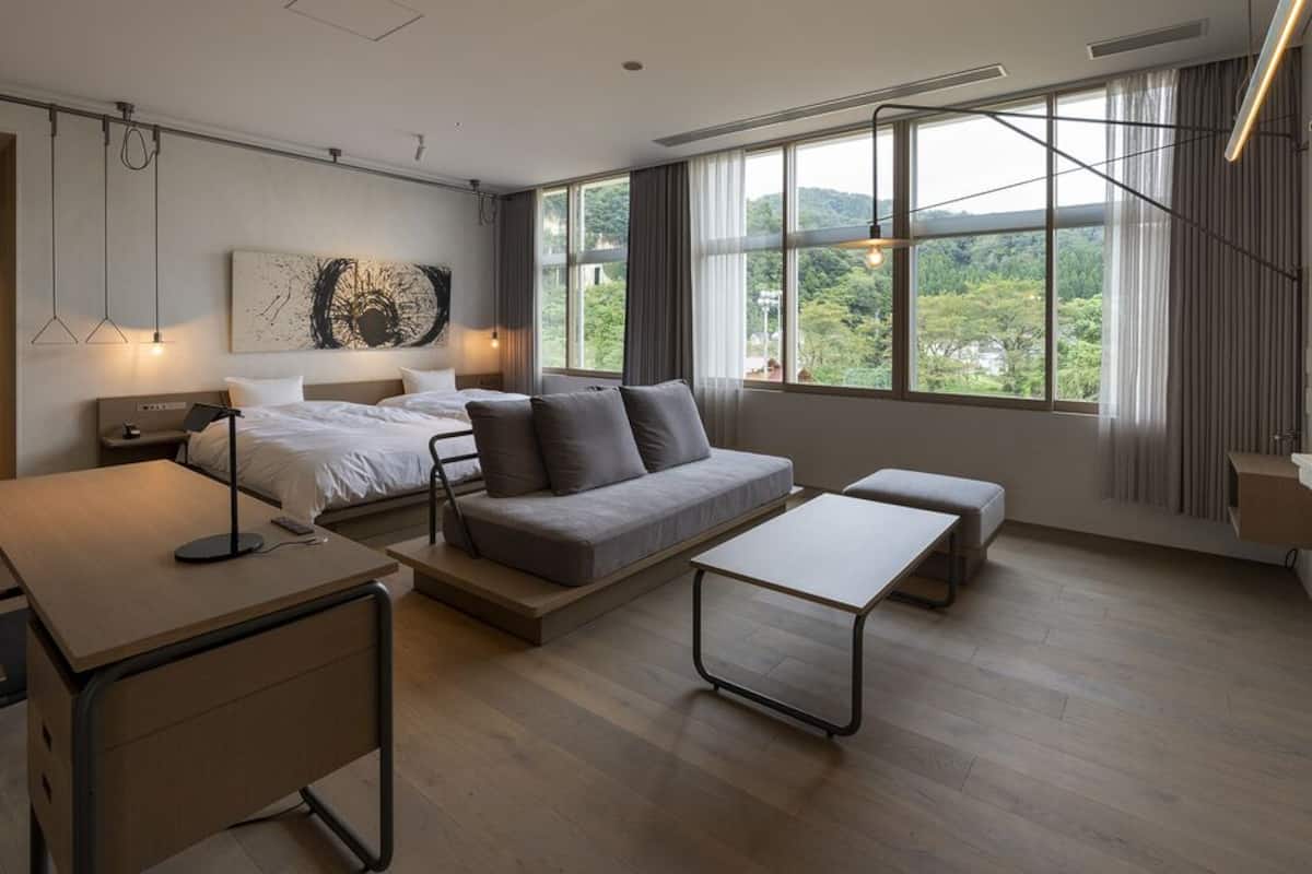 Een moderne hotelkamer met een bed, een bank, een bureau en grote ramen met uitzicht op groen.