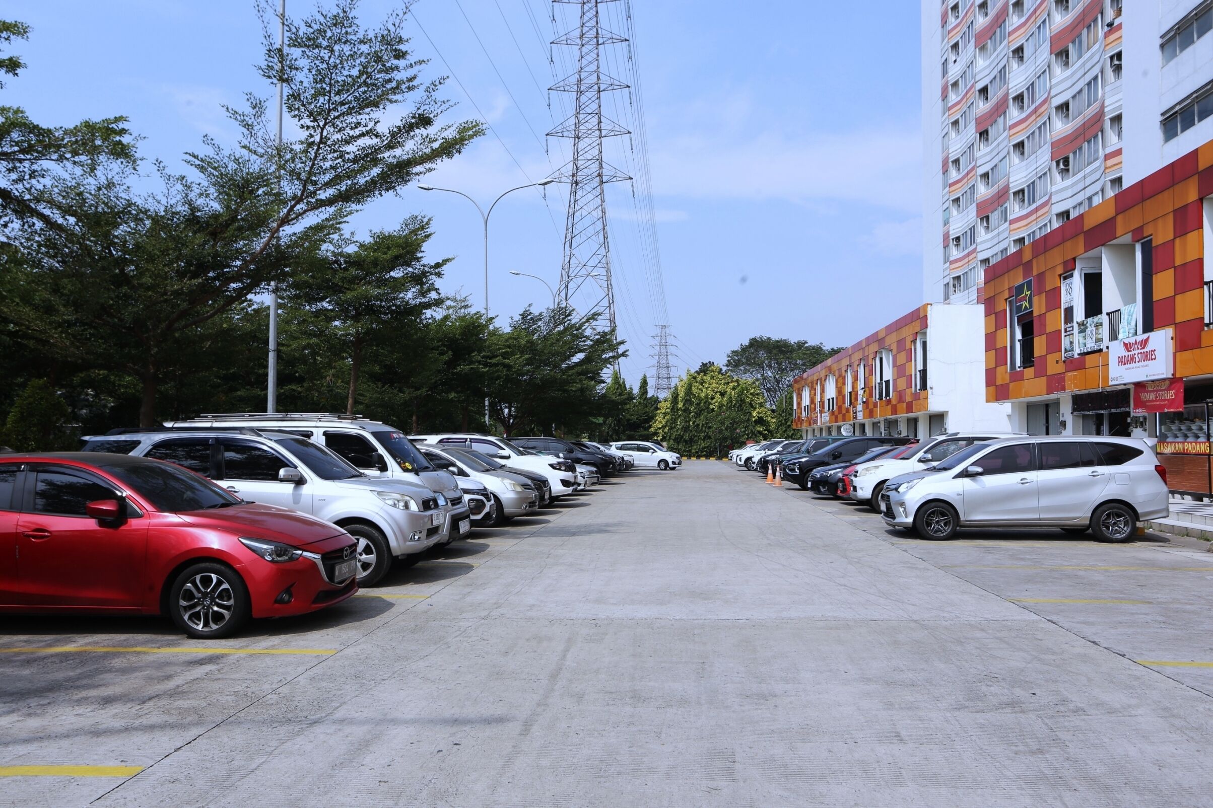 Un parking où de nombreuses voitures sont garées en rangées, un grand immeuble à la façade à motifs en damier sur la droite, et des arbres en arrière-plan.